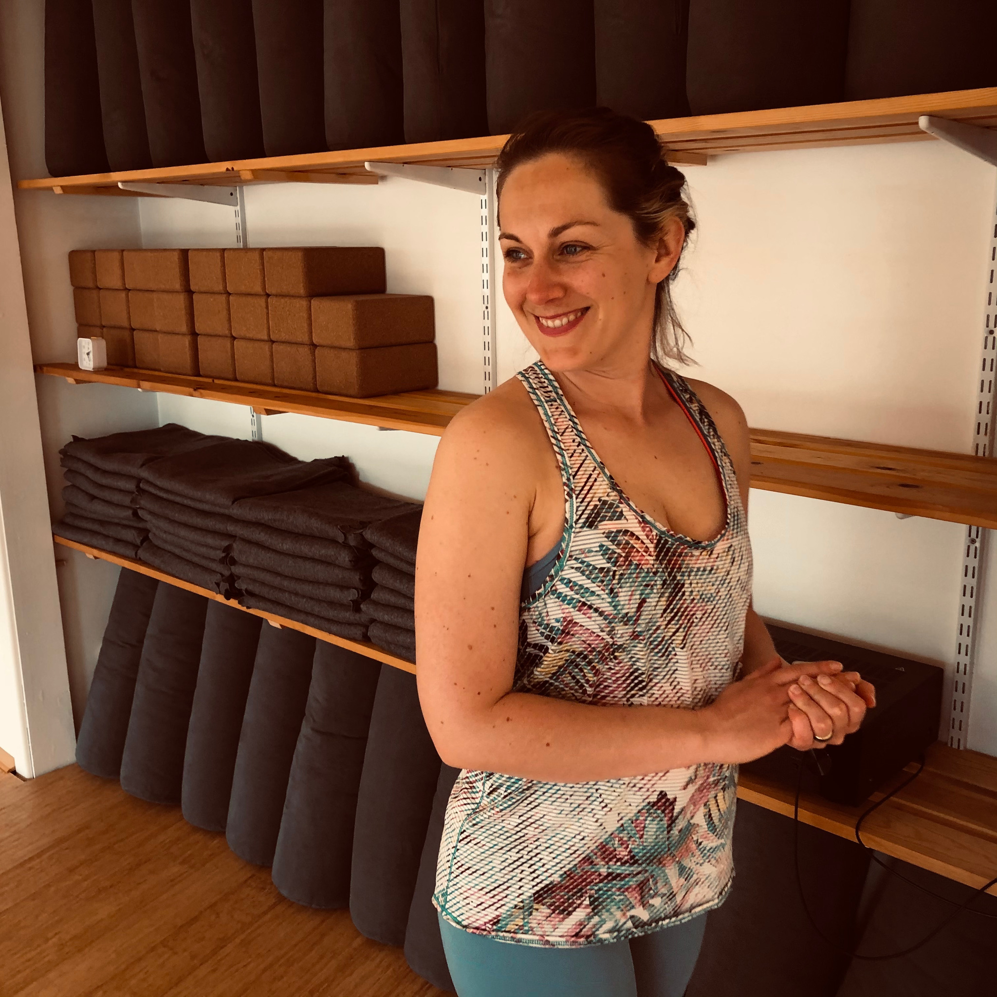 Smiling yoga teacher standing in front of a wall of yoga props in Yoga One Bournemouth.